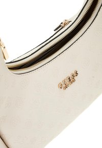 Beige handtas met textuur en een gegraveerd patroon, gouden hardware, dubbele ritsen en het "GUESS"-logo duidelijk zichtbaar aan de zijkant.