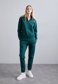 Σκούρο πράσινο φούτερ με φερμουάρ κατασκευασμένο από μαλακό ύφασμα, με ριχτές μανσέτες και ελαστική μέση, συνδυασμένο με λευκά αθλητικά παπούτσια.