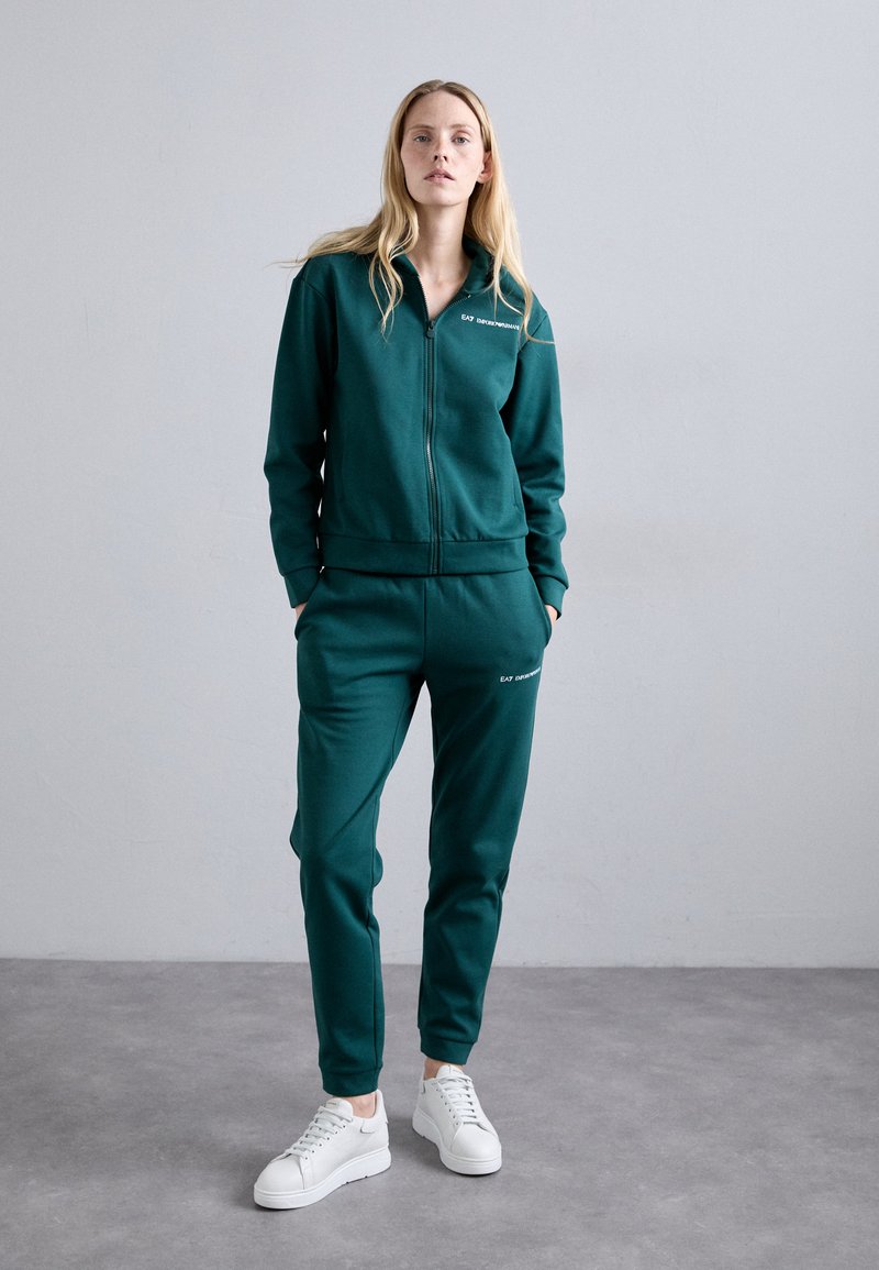 Σκούρο πράσινο φούτερ με φερμουάρ κατασκευασμένο από μαλακό ύφασμα, με ριχτές μανσέτες και ελαστική μέση, συνδυασμένο με λευκά αθλητικά παπούτσια.