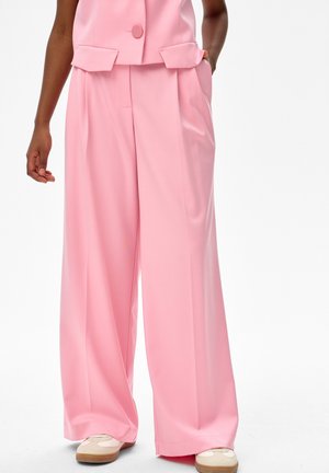 Osoba ubrana w różowe spodnie z wysokim stanem i szerokimi nogawkami oraz pasujący do nich różowy top bez rękawów, w zestawieniu z beżowo-białymi trampkami.