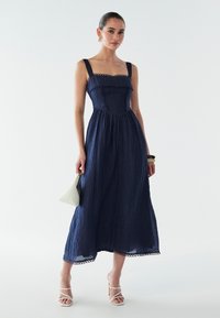 Vestido azul marino texturizado con escote cuadrado, costuras decorativas y dobladillo con borde festoneado. Combina con sandalias blancas de tacón y un pequeño bolso blanco.