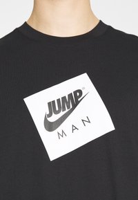 Černé bavlněné tričko s bílým obdélníkovým graphicem s textem "JUMP" a "MAN" v tučném černém písmu. Kolečkový výstřih.