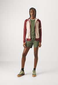 Rot und rosa Reißverschlussjacke über einem grünen Shirt und Shorts, mit einem schlanken Design, glattem Stoff und strukturierten Sneakers mit orangefarbenen Akzenten.