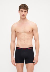 Caleçons noirs en tissu lisse avec un logo "HUGO" rouge sur la ceinture élastique. Design ajusté, coutures plates et taille mi-haute.