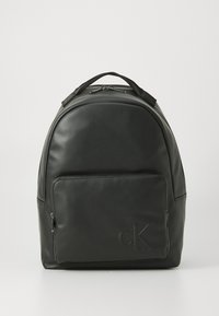 Mochila de cuero negra con bolsillo frontal con cremallera y logo "cK" en relieve, asa superior y cierre de doble cremallera sobre un fondo blanco.