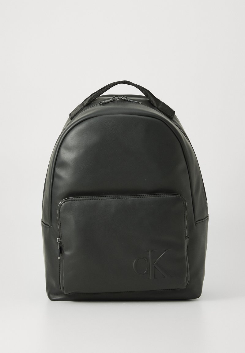 Mochila de cuero negra con bolsillo frontal con cremallera y logo "cK" en relieve, asa superior y cierre de doble cremallera sobre un fondo blanco.