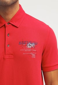 Homme portant un polo rouge avec une patte de boutonnage à trois boutons et un logo brodé bleu et blanc avec du texte sur la poitrine gauche.