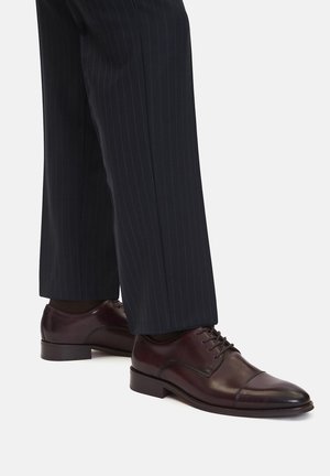 Brązowe skórzane buty wizytowe z zaokrąglonym noskiem i pięcioma dziurkami na sznurówki, zestawione z ciemnymi spodniami w cienkie prążki.