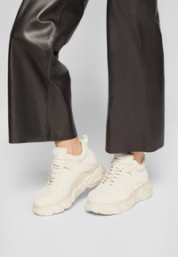 Crème-kleurige sneakers met een chunky zool, een gladde textuur, vetersluiting en een klein logodetail nabij de hak, gecombineerd met zwarte leren broek.