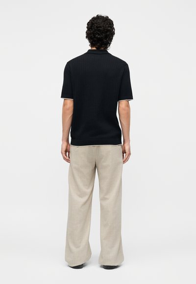 Άνδρας με σγουρά μαλλιά που φοράει μαύρο πόλο με κοντά μανίκια και μπεζ παντελόνια με φαρδιά μπατζάκια, στέκεται με την πλάτη στραμμένη σε απλό φόντο.
