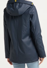 Marinblå vattentät jacka med huva och knappar. Har en slät textur och en avslappnad passform. Bakvy visar sömmar och designlinjer.