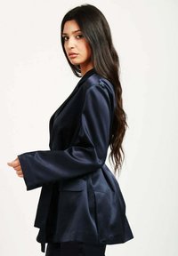 Blazer navy realizzato in tessuto liscio, con vestibilità aderente, maniche lunghe, due tasche frontali e un design del bavero classico.