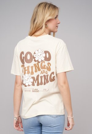 T-shirt en coton couleur crème avec un graphisme de style rétro comportant le texte "De bonnes choses arrivent" et des motifs de fleurs souriantes dans des tons bruns.