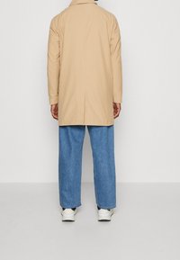 Persona vestita con un cappotto beige, jeans blu larghi e sneakers bianche, in piedi di fronte a un muro bianco semplice.