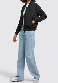 Černá bomber bunda se zipem vpředu, žebrovanými manžetami a dvěma bočními kapsami, v kombinaci se světle modrými širokými džíny a bílými teniskami.