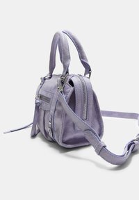 Petit sac à main en daim lavande avec deux courtes poignées, une poche zippée à l'avant, des accessoires en argent et une bandoulière longue réglable.