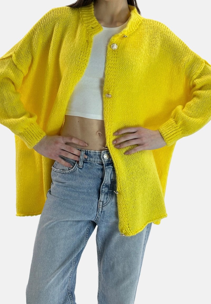 Personne portant un cardigan tricoté jaune vif ouvert sur un crop top blanc et un jean bleu taille haute, les mains posées sur les hanches.