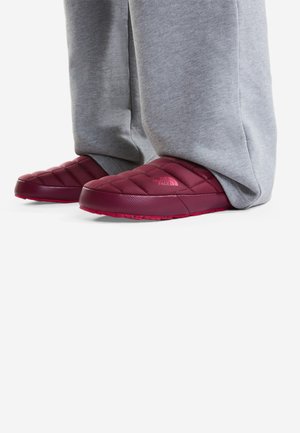 Person iført grå joggingbukser og mørkerøde vatterede slip-on sko med The North Face logo synligt på siden.