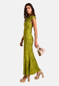 Grünes floralbedrucktes Abendkleid mit figurbetontem Schnitt, kurzen Ärmeln und ausgestellt geschnittenem Saum. Kombiniert mit goldenen Plateau-Highheels und einer beigen Clutch.