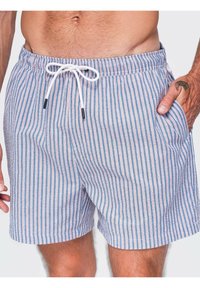 Torse masculin portant un short de bain bleu et blanc à rayures verticales avec un cordon blanc, une main dans la poche montrant un tatouage au poignet.