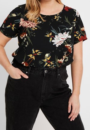 Vrouw draagt een zwarte, gebloemde korte mouwen blouse, ingestopt in hoge zwarte denim jeans met één hand in de zak.