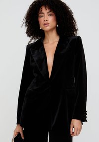 Schwarzer Samtblazer mit tiefem Ausschnitt, spitzen Revers und zwei Vorderseitentaschen. Verziert mit Knopfakzenten an den Ärmeln.