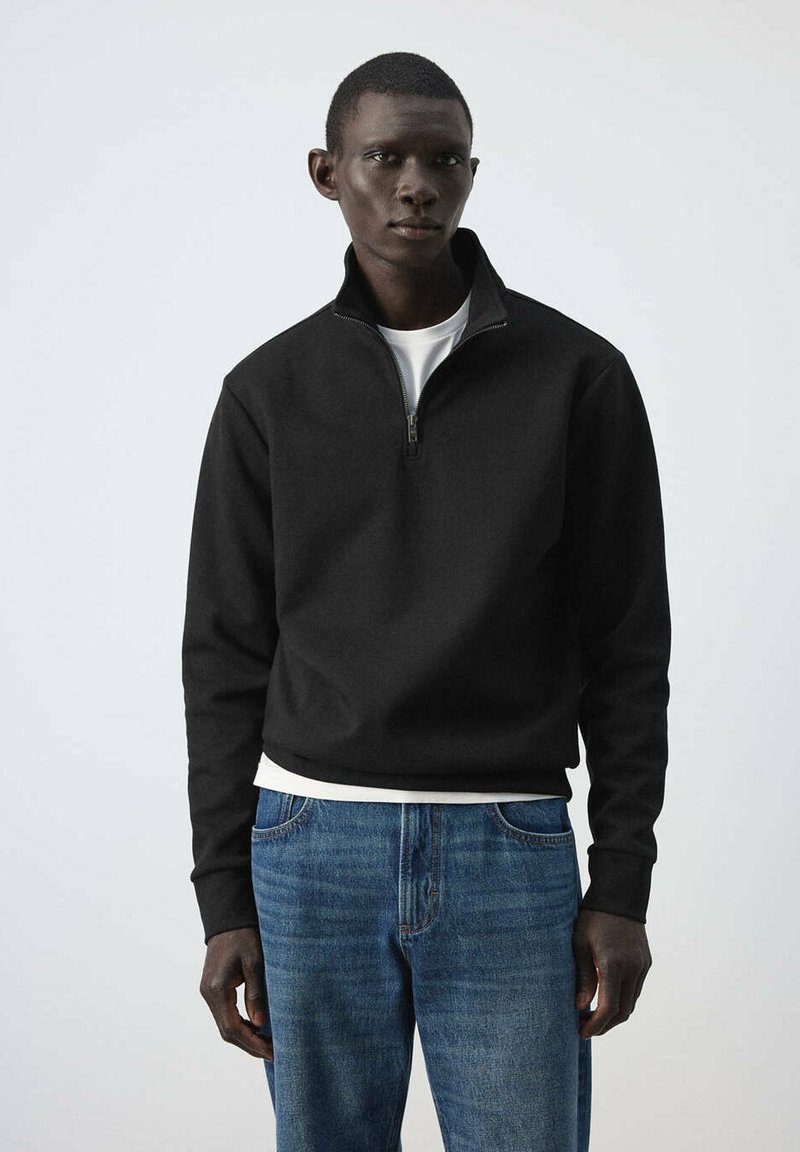 Homme portant un pull noir à fermeture éclair quart de zip sur une chemise blanche, associé à un jean bleu, debout devant un fond clair uni.