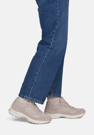 Person trägt beige Sneaker und hellbeige Socken mit blauen Jeans vor einem einfarbigen Hintergrund.