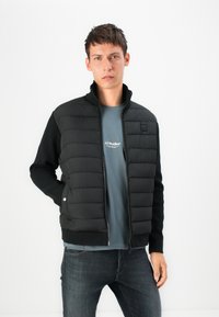 Black quilted jacket with a stand collar, zip front, and knit sleeves. Worn over a gray t-shirt and dark jeans, against a white background.