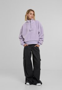 Paarse fleece pullover met een hoge kraag, rits en geborduurd logo; gecombineerd met losse zwarte cargobroek en witte sneakers.