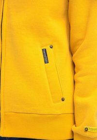 Felpa gialla realizzata in tessuto morbido, con chiusura a zip, tasca applicata e etichetta con marchio vicino al polsino. Superficie texturizzata.