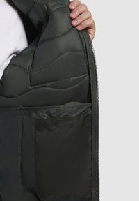Schwarze Pufferjacke mit gestepptem Innenfutter, ausgestattet mit einer Seitentasche. Sichtbarer Reißverschluss und glattes, glänzendes Material.