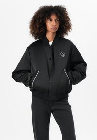Zwarte bomberjack met een ribbelkraag en boorden, voorzien van witte piping en een hartvormige smiley-embleem op de borst.