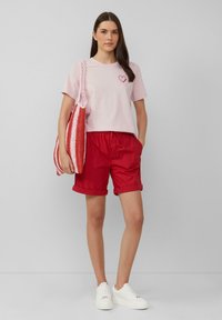 Roze katoenen t-shirt met rode hartborduurwerk, rode shorts met opgerolde mouwen en een gebreide rood-wit boodschappentas. Witte sneakers.