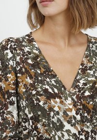 Ziedu raksta blūze zemes toņos - zaļā, brūnā un krēmkrāsā; ar V veida izgriezumu un teksturētu audumu, ar saplacinātām piedurknēm.