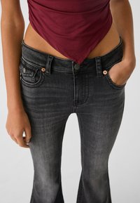 Jeans de mezclilla negros acampanados con cintura media, que presentan acentos de botones de cobre y bolsillos, combinados con una blusa ajustada en color burdeos.