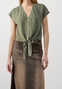 Grüne Bluse mit kurzen Ärmeln, V-Ausschnitt und Knopfleiste, am Bund gebunden, kombiniert mit einem braunen, satinartigen Midirock mit seitlichem Schlitz.