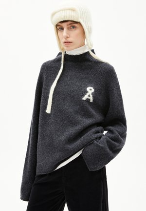 Grauer Wollpullover mit hohem Kragen, mit weißem gesticktem Logo. Das Modell trägt außerdem einen cremefarbenen Strickhut mit Ohrenklappen und Bändern.