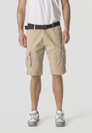 Hombre vistiendo pantalones cortos cargo beige con múltiples bolsillos, cinturón negro, zapatillas blancas y camiseta blanca, de pie frente a un fondo liso.