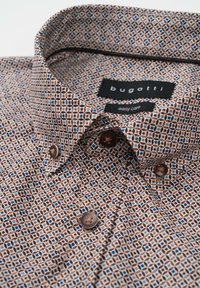 Brown patterned shirt with a geometric design featuring blue accents. Made of fabric with a smooth texture. Two brown buttons at the collar.