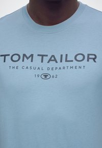 Κοντινό πλάνο ανοιχτό μπλε μπλούζας με μαύρο κείμενο "Tom Tailor The Casual Department 1962" στο μπροστινό μέρος που φοριέται από άτομο.