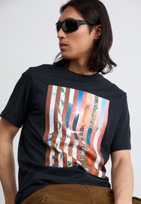 En person med langt hår bærer solbriller og en sort T-shirt med et farverigt, stribet grafisk design, sat mod en neutral baggrund.