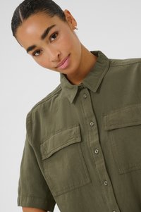 Jeune femme avec des cheveux noirs lisses portant une chemise boutonnée olive avec des poches à rabat et une petite boucle d'oreille en argent, souriant doucement.