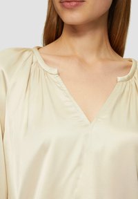 Blouse en satin beige avec un décolleté en V et des épaules froncées, présentant une texture lisse et un drapé doux autour du cou.