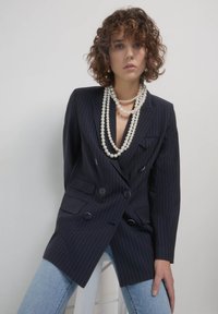 Blazer doppiopetto navy a righe sottili con rever a contrasto, grandi bottoni neri e tasche frontali, indossato sopra jeans azzurri chiari e con perle sovrapposte.