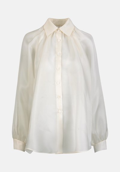 Krēmkrāsas, garām piedurknēm blūze ar pogu aizdari priekšpusē, smailu apkakli, savāktām plecu daļām un pogām apsienamiem aproču galiem.