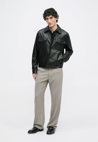 Μαύρο δερμάτινο μπουφάν με κουμπιά, δύο τσέπες στο στήθος και κλασικό γιακά, συνδυασμένο με ανοιχτό γκρι καρό παντελόνι και μαύρα παπούτσια.