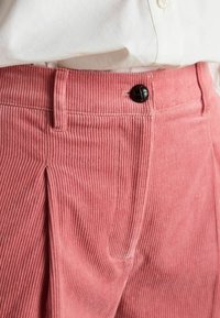 Rozā kordurojas bikses ar vertikālu ribojumu, kas ir ar vienu krokumu, melnu pogu aizdari un klasisku jostasvietas dizainu.
