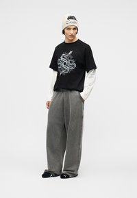 Νέος άνδρας που φοράει λευκό σκουφάκι, μαύρο T-shirt με σχέδιο πάνω από λευκά μακριά μανίκια, χαλαρό γκρι παντελόνι και μαύρες παντόφλες Puma, στέκεται με τα χέρια στις τσέπες.