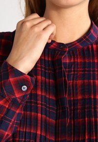 Chemise à carreaux rouge et bleu marine, avec un col arrondi, un devant plissé et des manches retroussées avec des détails de boutons. Texture de tissu lisse.